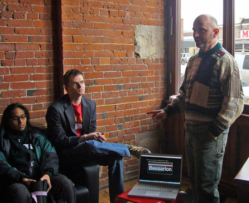 Me in sweater with laptop on stool and two people perched on a nearby sofa