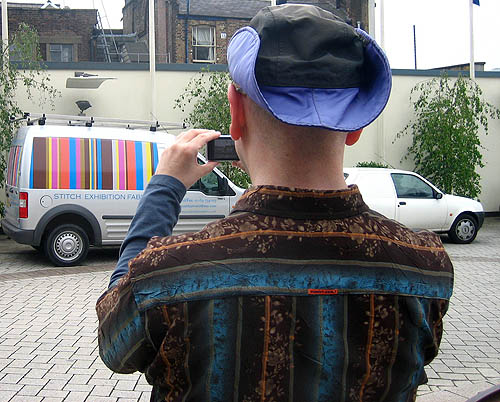 From behind, I take a picture of a van with coloured stripes on the side while wearing a patterned shirt, a purple-and black hat