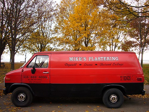 By a park whose trees have bright yellow and green leaves, a red van with black bottom panels is labelled MIKE’S PLASTERING, with Drywall • Stucco • Textured Ceilings in script