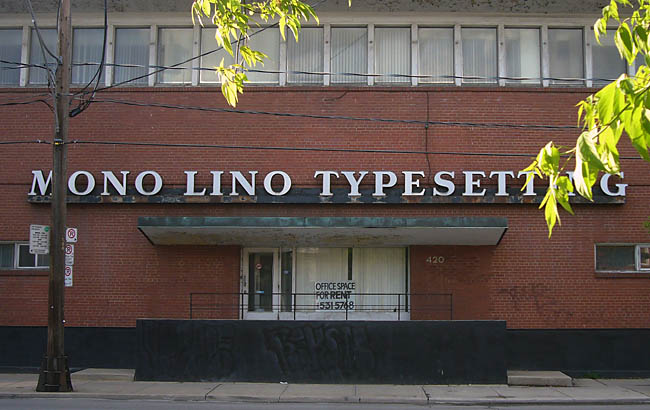 Behind a few green-leafed branhes, a hulking building has a sign reading MONO LINO TYPESETTING