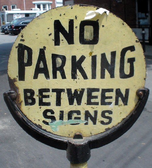 Round sign with thick black metal base reads NO PARKING BETWEEN SIGNS, with PARKING scooped into an arc in the middle