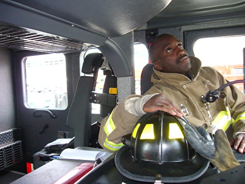 Black fireman inside cab of large fire apparatus, with helmet nearby