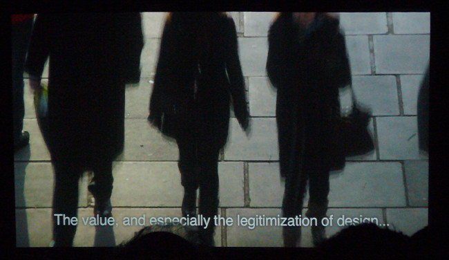 People’s heads partially block the view of a film frame showing people walking down a cobbled street. Subtitle: The value, and especially the legitimization, of design