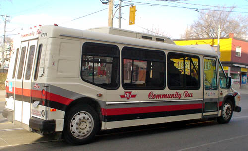 Long white shuttle bus with grey, red, and black stripes, windows half the height of the bus, and a single door behind a large front wheel is emblazoned Community Bus in script type