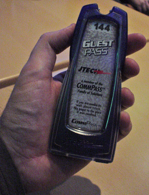 Narrow, palm-sized plastic pager numbered 144 reads ‘Guest Pass JTech... If you are unable to wait, please return this pager to the place it was received. CommPass™’