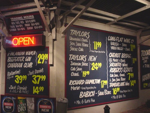 Hand-drawn sign wraps around corner of bottle shop, with white writing on jet-black background and a neon sign reading ‘OPEN’
