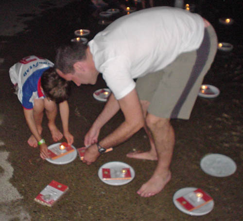 A man and a boy lean over and move two books and a candle on a floating paper plate