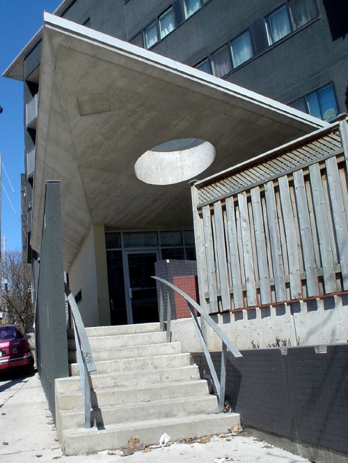 Black building angles away from entrance, with concrete steps, curven metal banister, and circular hole cut out of triangular angled roof