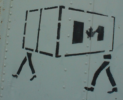 Stenciled sign on metal siding shows a cargo container, emblazoned with a Canadian flag, that walks away on two pairs of legs