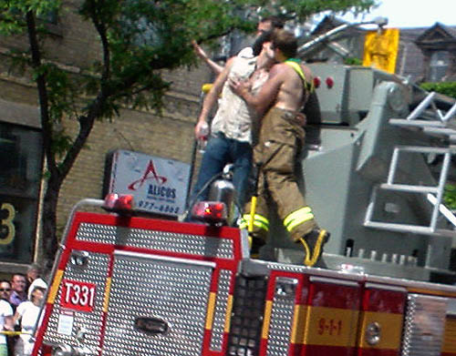 Shirtless fireman in suspenders and turnout pants plants a kiss on man in cowboy hat atop a firetruck