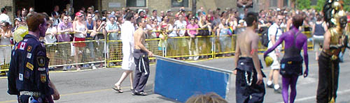 In a cordoned-off parade, a red-haired man in blue overalls covered with patches follows two shirtless guys holding a blue banner, while others wear purple body paint or a gold headdress