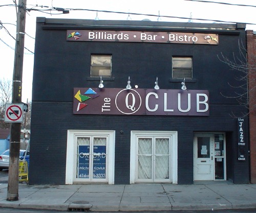 Black building has signs reading ‘Q Club’ and ‘Billiards · Bar· Bistro.’ A for-lease sign sits inside a door