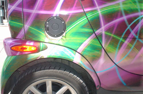 Close-up shows a Smart Fourtwo’s black fuel-filler cap, red reflector, and black tire amid a paint job of bright red, green, pink, and blue arcs and curls