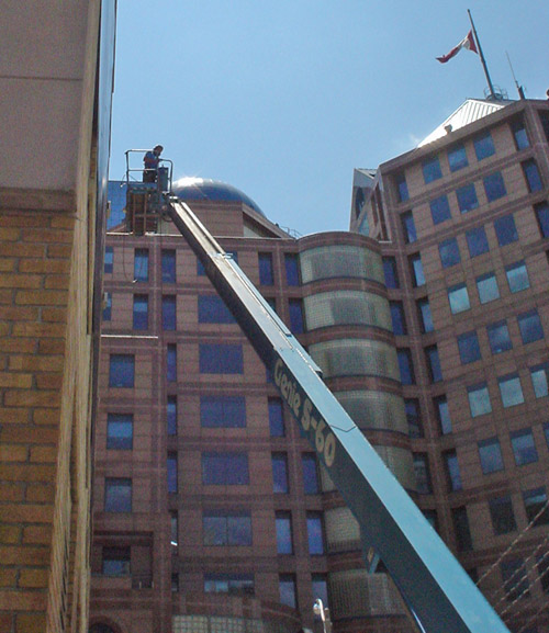 Man stands in gondola in midair on the end of a huge blue metal arm (labeled Genis S-60) an arm’s reach from a brick building