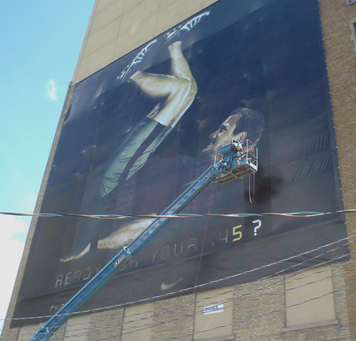 Long metal arm holds metal gondola close to a three-storey banner showing a man in T-shirt and Lycra shorts pressing his feetup to the top edge