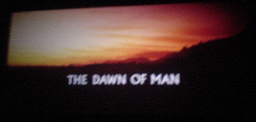 Frame from film shows sunrise on a savannah and the title THE DAWN OF MAN