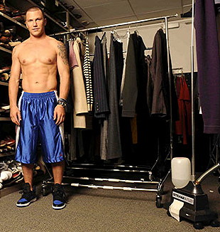 Shirtless Sean Avery, wearing excessively long blue nylon shorts and standing near a clothes rack on wheels