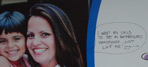Billboard shows woman holding young boy. Graffiti alongside draws in a dialogue balloon reading ‘I want my child to be a repressed homophobe just like me’