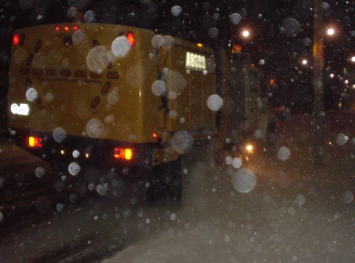 Yellow truck body drives through away through flake-strewn image alongside a white-covered sidewalk