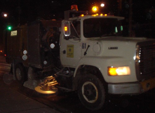 White truck has an illuminated brush and vacuum attachment behind the cab at ground level