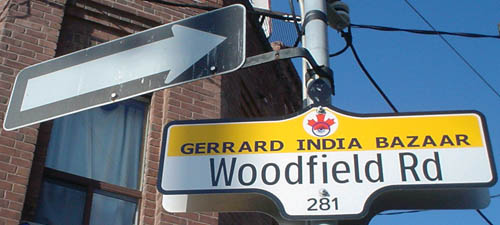 One-way arrow stands on same pole as sign reading Woodfield Rd. in Clearview and GERRARD INDIA BAZAAR in Verdana