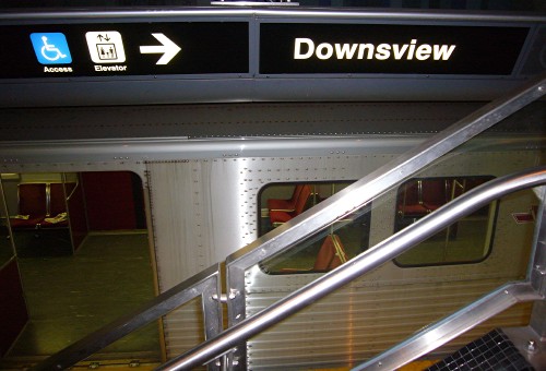 Sign over parked subway car has two pictographs, an arrow, and the word Downsview