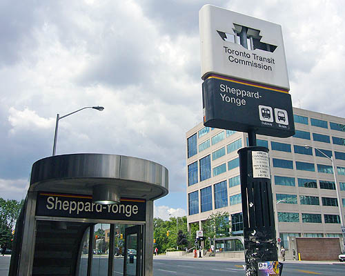 Entrance has massive semicircular steel canopy and reads Sheppard-Yonge. Nearby standing sign has missing TTC logos, through which clouds are visible