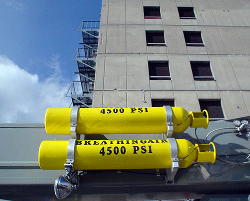 Yellow pressurized canisters labelled BREATHING AIR 4500 PSI are attached to metal surface as bland concrete building with fire escapes looms behind