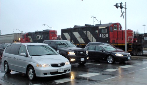 Four lanes of vehicle traffic are stopped at a line alongside two locomotives and a railcar