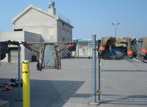 Chain-link fence is draped with firefighter turnout coats, one of them spread-armed against the mesh