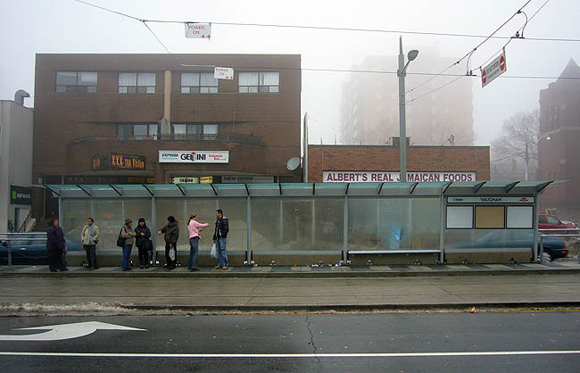 Seven people wait in the fog under a half-block-long glass shelter in front of ALBERT’S REAL JAMAICAN FOODS