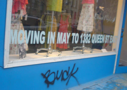 Words (in Arial) plastered on bottom of dress-store window read MOVING IN MAY TO 1382 QUEEN ST EAST (with graffiti on wall below)
