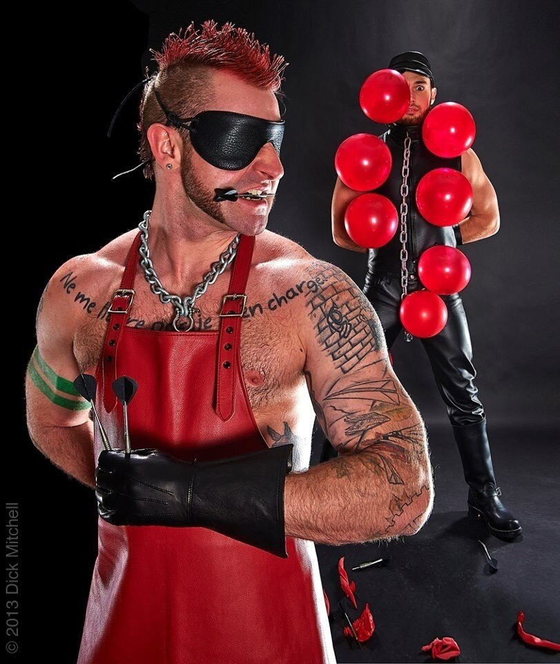 Man in red mohawk wears a red leather butcher’s apron and a blindfold. Holding darts in his hand (and one between his teeth), he almost looks around to see another man in a leather suit and chain who is festooned with balloons and wearing an anxious expression