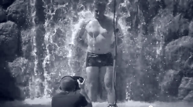 Embarrassed-looking bobsledder in square-cut swimsuit stands under waterfall as male photographer takes his picture