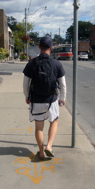 Seen from behind, a man in shorts, backpack, and baseball cap walks down the street