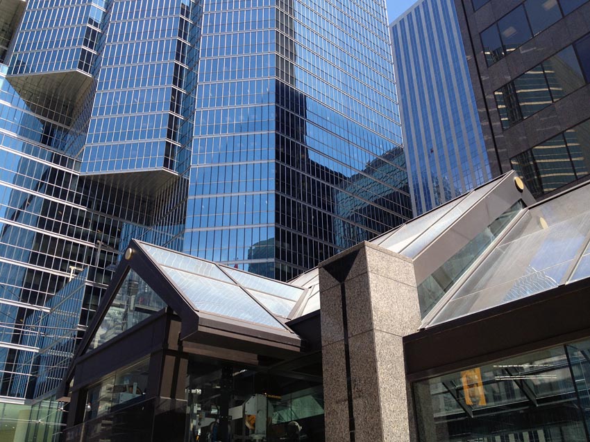 A-frame roofs in glass and granite columns with triangular incisions at top stand before soaring skyscrapers