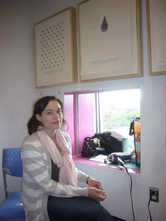 Amanda seated at window under posters (with hair dryer nearby)