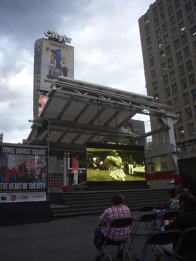 Man in giant lamé dress on video screen in open square