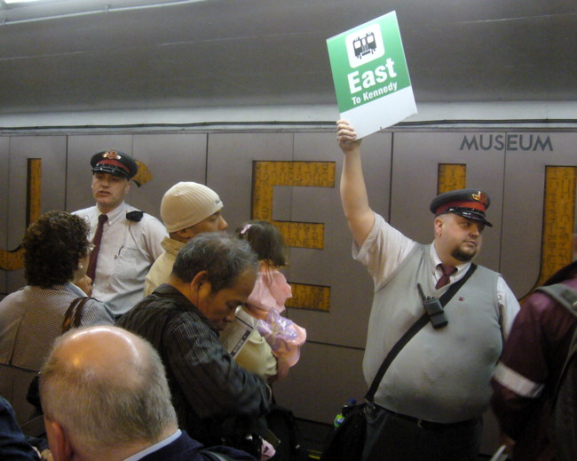 Supervisor in cap holds up green sign reading East To Kennedy