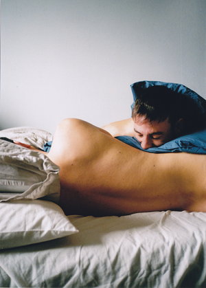 Man leans over and kisses flank of other man through blue bedsheet