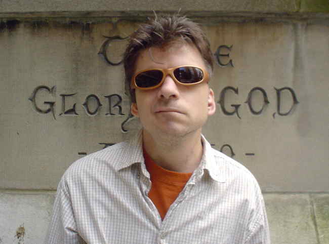 Man in orange-rimmed shades stands before stone inscribed TO THE GLORY OF GOD