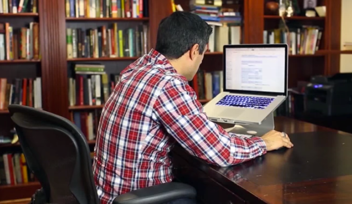 Man in plaid shirt hunched over desk working on a MacBook on a stand