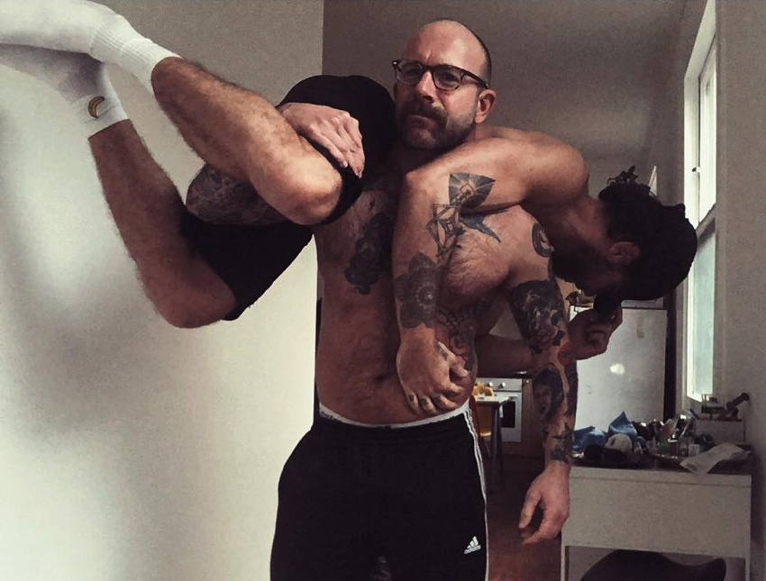Tattooed shirtless male holds another shirtless male over his shoulders as if a sack of potatoes