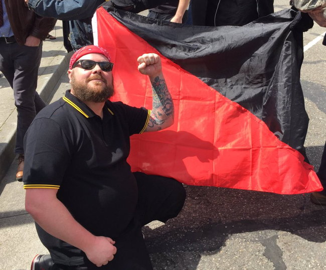 Bearded man in Fred Perry shirt holds up fist (at end of tattooed arm) in front of flag divided diagonally into black and red halves