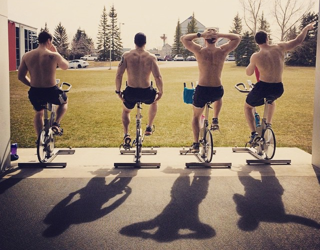 Four shirtless men on exercise bikes at a garage-door ramp