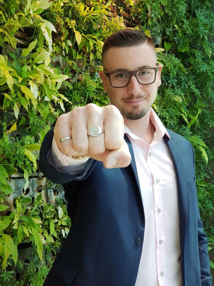 Alex Kopacz, in a suit jacket, a tidy haircut, and glasses, holds his fist to the camera, showing iron ring and Olympic ring