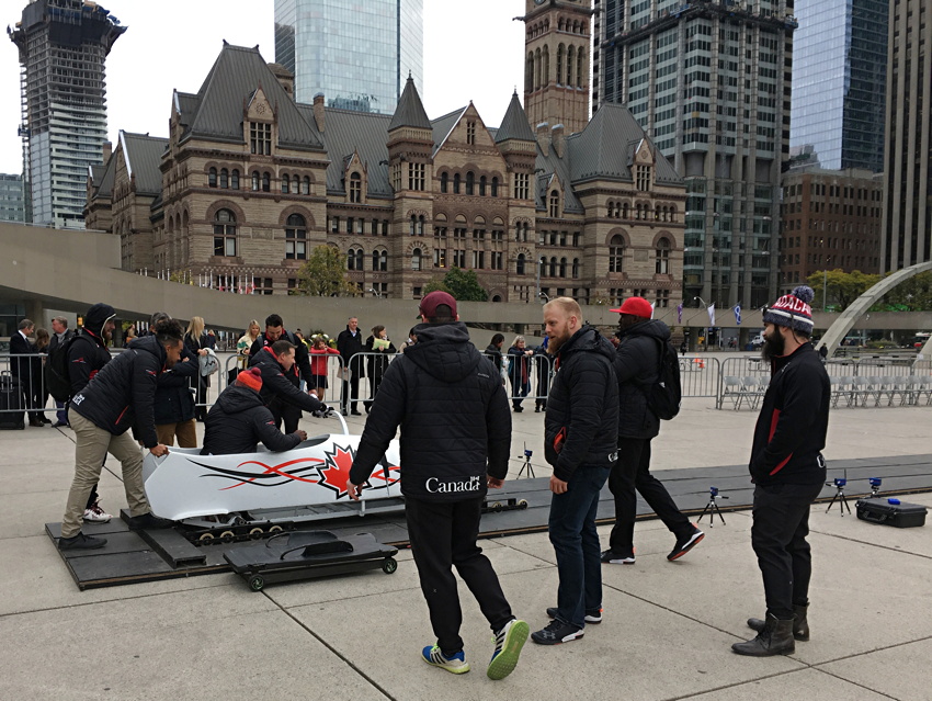 Dudes in black uniforms emblazoned with the Canada wordmark futz around a white bobsled