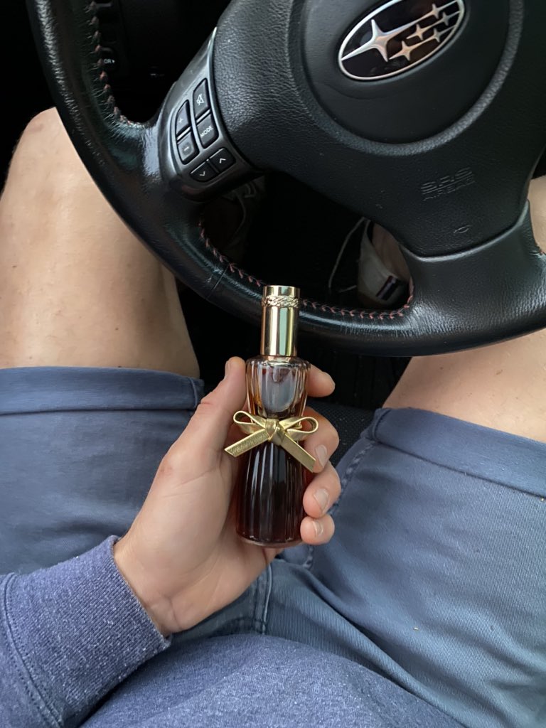 Gloved hand at steering wheel holding deep-red perfume bottle with gold bow