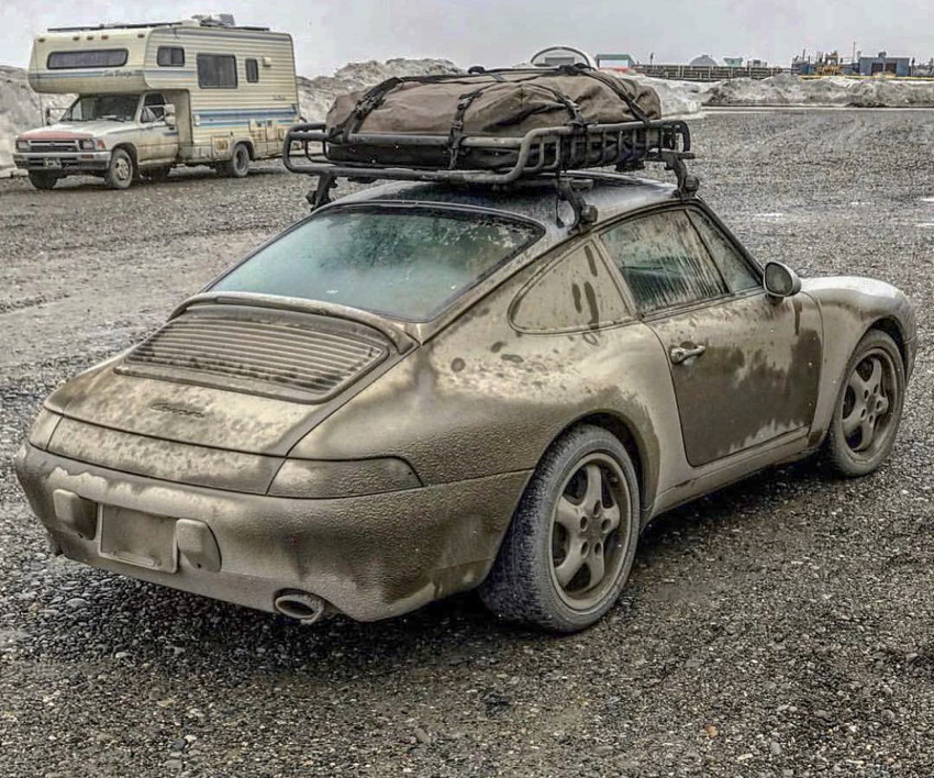 Porsche 911, in a gravel field, is coated with black dust, and has tarp-enclosed cargo on a roof rack