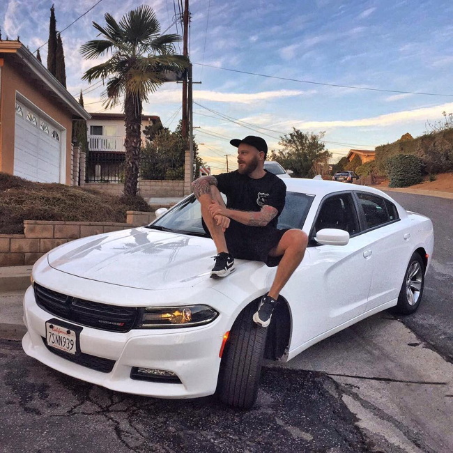 At sunset on a street lined with palm trees, man in black shorts, T‑shirt, and ballcap sits on hood of white Dodge Challenger, left foot on out-turned tire